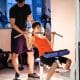 foto horizontal de uma pessoa adulta iniciante recebendo orientação de um professor em uma máquina de musculação. O ambiente pode ter outros frequentadores ao fundo